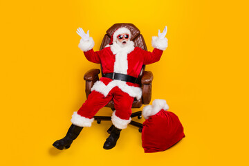 Fototapeta na wymiar Full size portrait of eccentric impressed santa sit chair raise arms new year presents bag isolated on yellow color background
