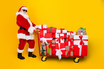 Full body profile portrait of eccentric santa push market trolley pile stack christmas giftbox isolated on yellow color background