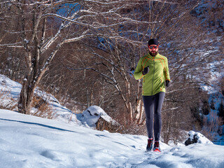 Fototapeta na wymiar A bearded man is jogging outdoors high in the mountains in winter. A guy in a green jacket and sunglasses is training. Trail running