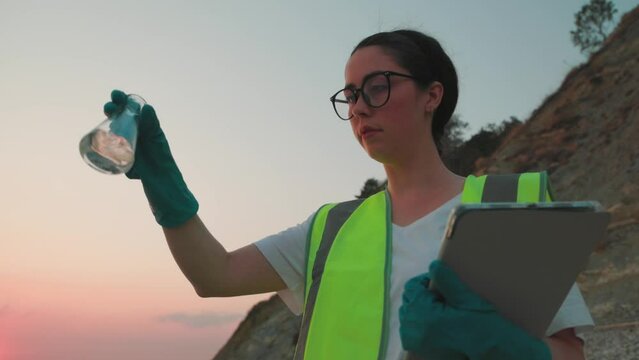 Bottom view of young woman ecologist in rubber gloves shakes flask with sample of water from ocean and check this with tablet. Concept of ecology and recycle resources