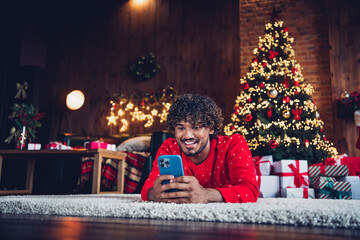 Photo of young happy funny guy lying floor using smartphone texting his girlfriend waiting at home new year atmosphere party indoors