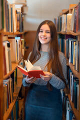 Vertical shot of smiling brunette student reading book for pleasure in the library. Getting...
