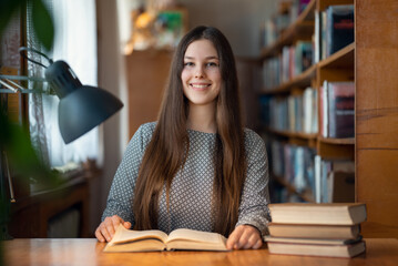 Female student sitting at the desk, reading a book and smiling. Brunette girl with long hair enjoys studying time at the library, getting education with pleasure