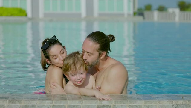 Happiness and harmony in family life. Happy family concept. Young mother and father kissing his little son. Happy family resting together in swimming pool on sunny day.