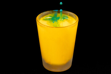 Copo de vidro cheio com bebida de cor amarela e gota de liquido de cor azul caindo. 