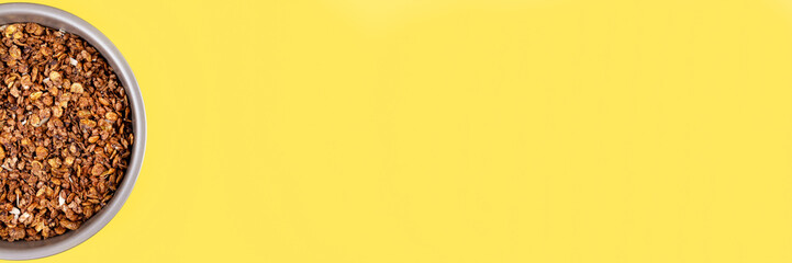 Roasted chocolate granola cereal in round baking tray on bright yellow background banner. Top view, flat lay muesli. Panoramic web header. Wide screen wallpaper
