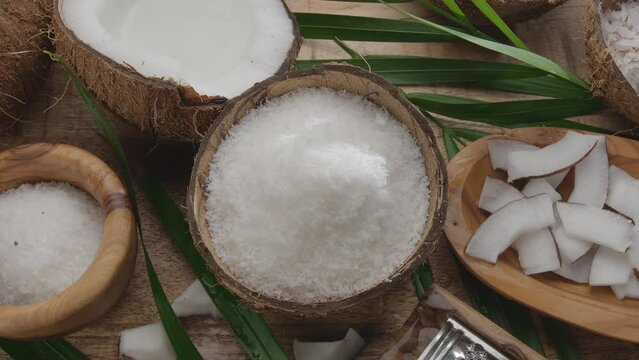 Fresh opened coconuts along with coconut flakes, slices and coconut leaves are slowly rotating on a wooden table.