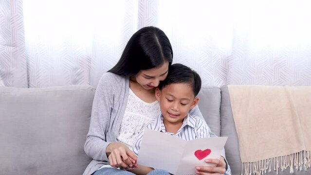 Asian woman hugging cute boy in arm reading Mother's day card DIY with tender love bonding relationship, preschool son happy spend time with mother in weekend leisure, single mom motherhood lifestyles