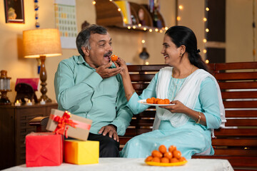 daughter and father eating sweets by sharing together at home during diwali festival celebration at home - concept of festive wishes, relationship bonding and holidays.
