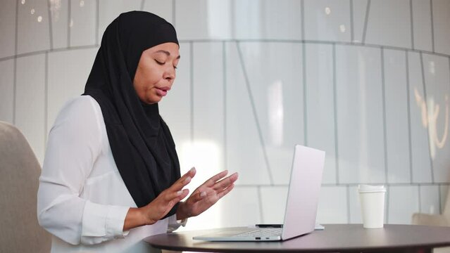 Anxious multicultural woman in black hijab having inexplicable fear and hand tremor during focused work on wireless laptop. Mature lady suffering from panic attack at modern corporate workspace.