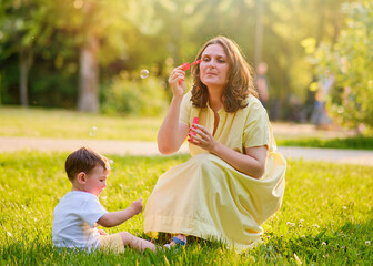 A mother woman plays with a baby in nature blowing soap bubbles. Happy child and mom play on the green grass in the summer park. Kid aged about two years (one year eleven months)