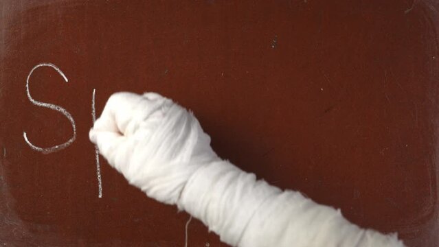 Textured brown chalkboard background. Mummy's hand in bandages writing 'spooky' on the board with a piece of white chalk in close up.