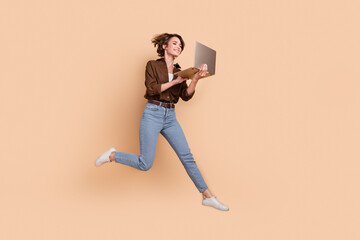 Fototapeta na wymiar Full length photo of excited pretty girl dressed brown shirt jumping high working modern gadget isolated beige color background