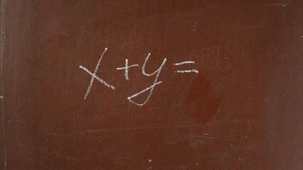 Textured brown chalkboard background. x and y addition equation is written on the board with a piece of white chalk. Close up shot.