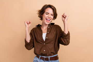 Photo of pretty excited girl dressed brown shirt screaming yes rising fists closed eyes isolated...