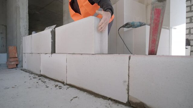 Bricklayer applies adhesive glue on autoclaved aerated concrete blocks with notched trowel. Brickwork worker contractor removes excess mortar from foam concrete doing masonry with laser level.