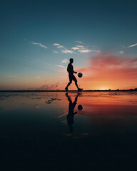 man juggles a soccer ball on the beach, water reflection, blue sky and red color sunset, outdoor...
