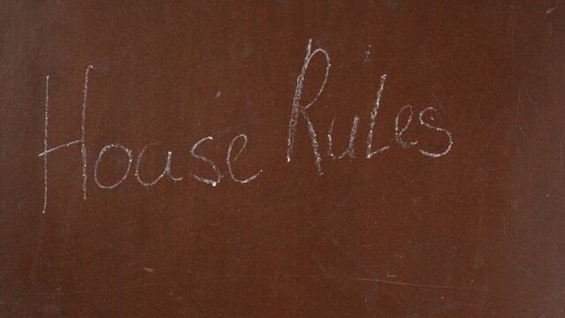 Textured brown chalkboard background. 'House rules' is written on the board with a piece of white chalk. Close up shot.