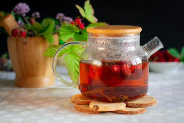 A glass teapot filled with bright reddish herbal tea stands on a wooden hot stand, behind a mortar...