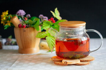 A glass teapot filled with bright reddish herbal tea stands on a wooden hot stand, behind a mortar with medicinal herbs and berries