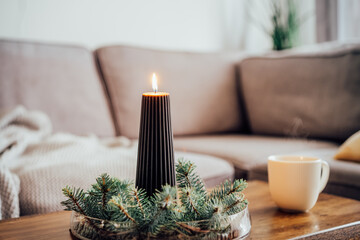Composition of flaming black christmas candle and spruce branches and cup with hot drink on coffee table in living room. Natural Christmas decorations for home. Cozy Christmas winter domestic mood.