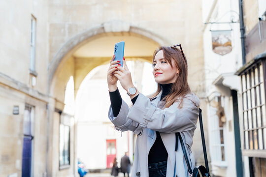 Young tourist woman visiting a picturesque city street destination, using smart phone taking pictures during holiday and travel walk in old European city, outdoors. Sightseeing trip through the city.