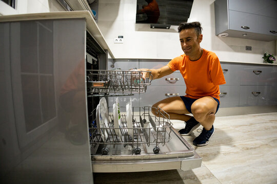 An adult man with an amputated arm is crouched in the kitchen of his home putting dishes in his sink. Concept of housework in disabled people.