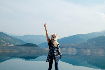 Female traveler wearing a broad brim hat enjoying beautiful view of a mountain lake. Young woman experiencing spiritual uplifting from being alone in nature. Background, copy space, back view.