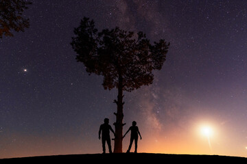 Couple in love standing on the hill near big tree, and looking at the Milky Way galaxy .