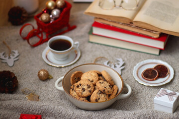 Bowl of cookies, cup of tea, dry oranges, pine cones, books, reading glasses, small presents, various Christmas decorations and lit candles on the table. Cozy Christmas hygge. Selective focus.