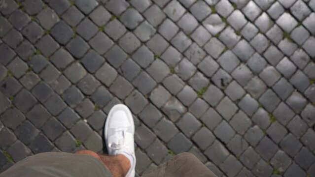 Closeup top view first person view of unrecognizable man wearing white sneakers walking down on paving stones on city street on bright sunny day, pov video. Shooting in slow motion.