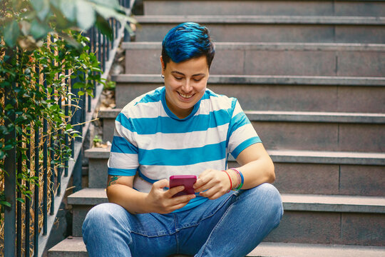 In a serene urban setting, an individual with striking blue hair and a striped shirt is engrossed in their phone. The casual yet engaging scene evokes the blend of nature and tech in modern life.