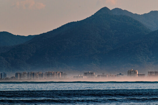 Bertioga city skyline, with its buildings covered by a fine mist, under a cloudless blue sky.