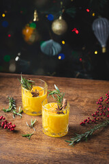 Orange winter cocktail with Christmas spices: star anise, cinnamon stick and rosemary on a background of Christmas tree and lights. Winter cocktails for warming up and winter holidays
