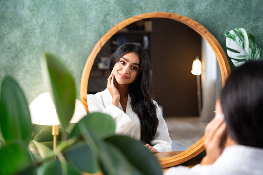 Hair care and self-care with beautiful Indian woman looking in mirror touching her healthy long hair sitting at the dressing table.