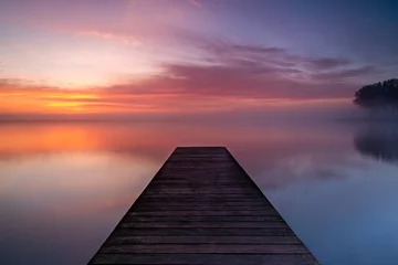 Fotobehang A wooden pier extends into the lake, where a layer of fog forms above the calm water. The rising sun beautifully colors the sky. © Bram