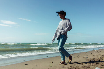 A beautiful brunette girl stands barefoot on the sandy seashore, enjoying the nature