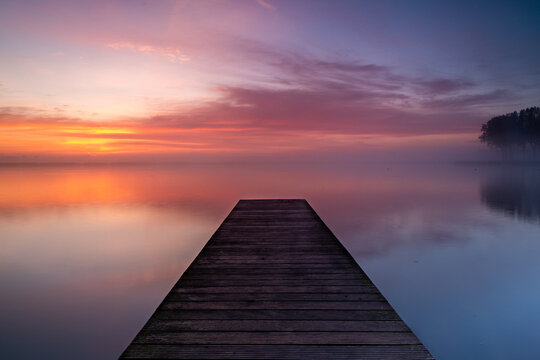 Fototapeta A wooden pier extends into the lake, where a layer of fog forms above the calm water. The rising sun beautifully colors the sky.
