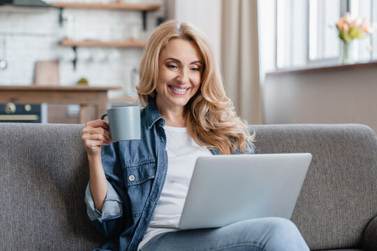 Happy relaxed mature woman resting on the sofa, watching movies films webinars video on laptop, drinking hot coffee tea beverage at home. Online shopping, remote learning - Powered by Adobe