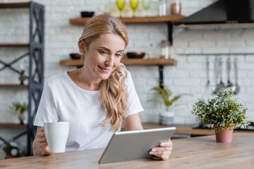 Happy mature Caucasian woman drinking hot coffee tea beverage, having breakfast at home while reading e-book, using tablet for watching movies videos webinars tutorials online at home kitchen
