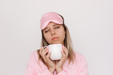 A young sleepy blonde woman in a pink pajamas and sleep mask with her eyes closed holding white cup...