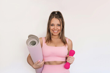 Young caucasian smiling fit blonde woman in pink sportswear holding yoga mat and dumbbell in her hands isolated on a white background. Fitness, workout concept. Healthy lifestyle, wellness, sports