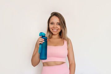 Young caucasian smiling fit blonde woman in pink sportswear holding sports blue water bottle in the hand isolated on white background. Fitness concept, healthy lifestyle