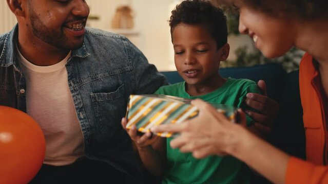 Close-up of father and mother surprising little son with a gift and balloons. African American dad and mom show their love and care to son