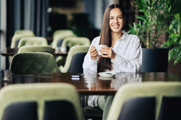 Portrait of a young woman sitting in a coffee shop and holding a cup of fresh morning coffee