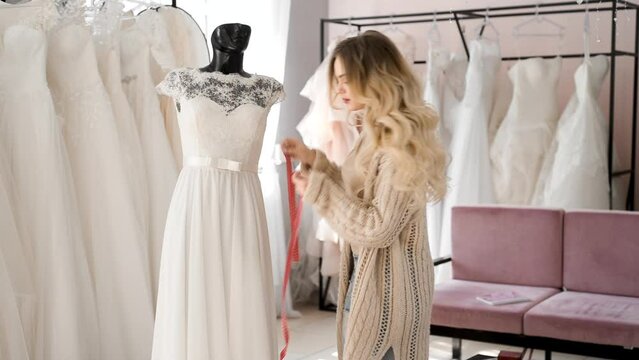 A young woman works as a seamstress in a wedding dress salon. In a wedding dress showroom, the sales consultant selects the dress and measures the length with a centimeter. Small business self-employe