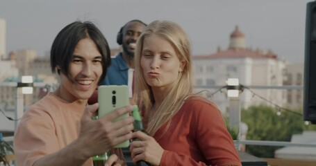 Asian man and Caucasian woman posing for smartphone camera taking selfie sharing photo at rooftop party. Modern technology and entertainment concept. - Powered by Adobe