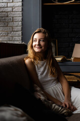 Fototapeta premium A young woman in a white dress sits on a sofa in a gloomy room and looks to the side. self-reflection, reflections