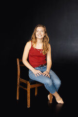 A young woman in blue jeans and a red T-shirt sits on a chair on a black background and smiles