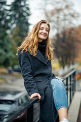 Obraz na płótnie Canvas A woman in a long gray coat and blue jeans on white sneakers sits on the railing and looks at the camera on the street of an autumn city